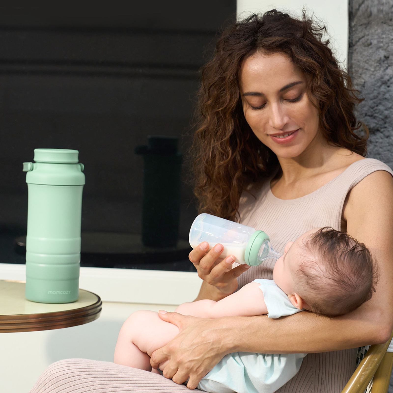 Une femme nourrit un bébé avec un biberon. Une bouteille verte est sur la table à côté d'une bouteille d'eau verte. Momcozy est écrit sur la bouteille d'eau.