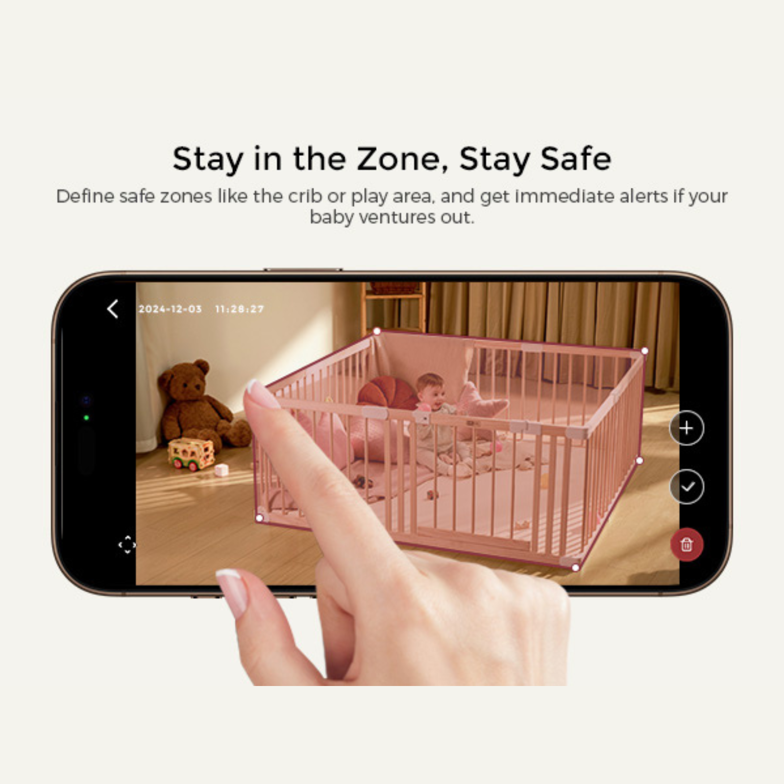 A phone displays a baby in a playpen. A hand touches the screen. Teddy bear and toys are visible.