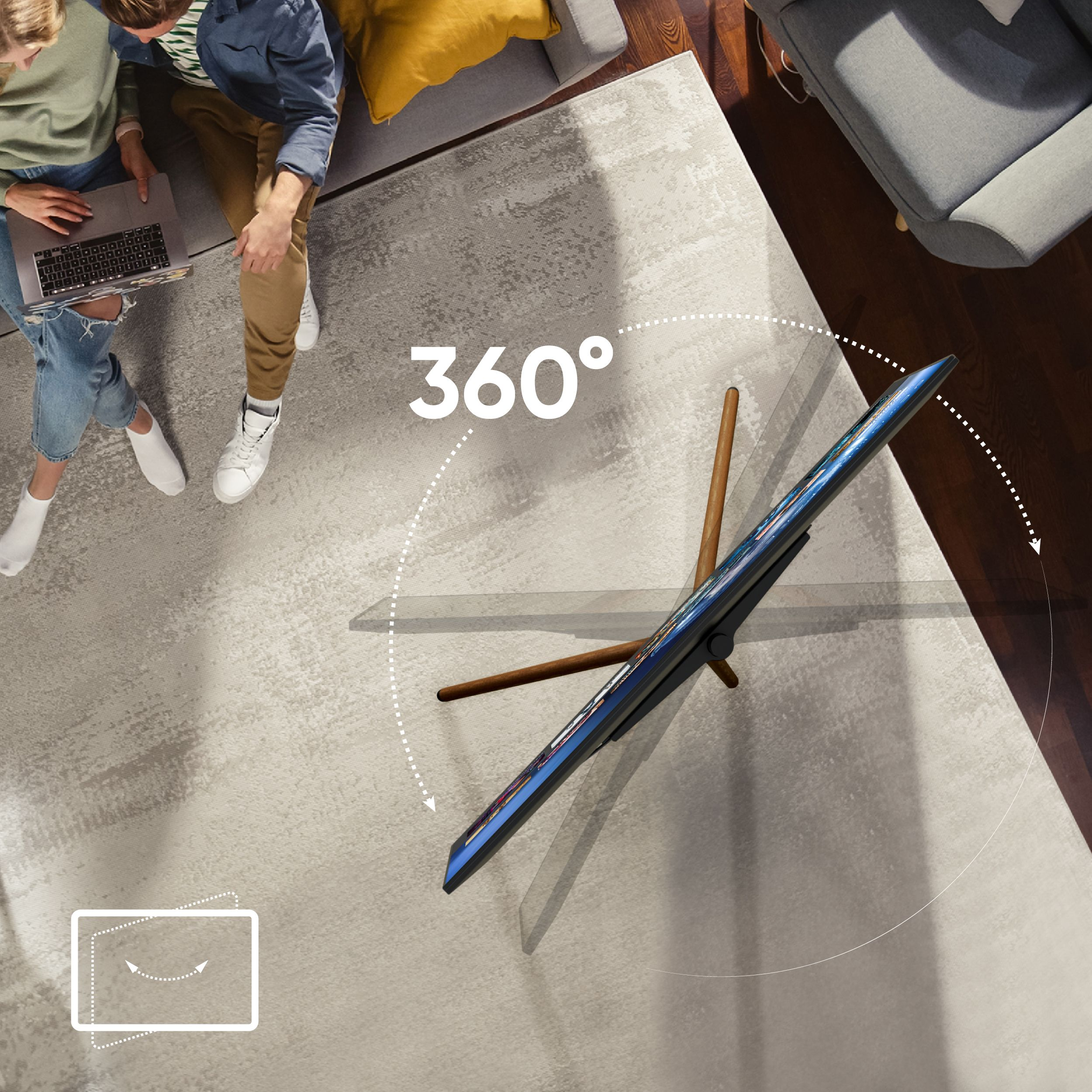 Dwie osoby siedzą na sofie z laptopem. Telewizor obracany o 360° jest wyświetlany na drewnianym stojaku.