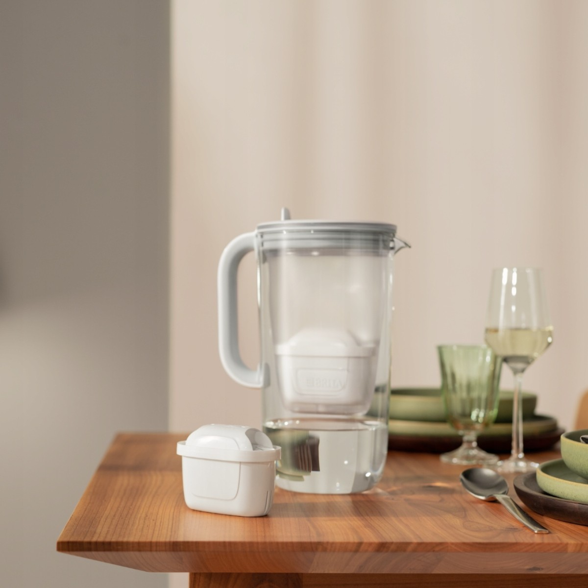 Dzbanek z filtrem wody z białym filtrem stoi na drewnianym stole. W tle widać kieliszek wina i naczynia.