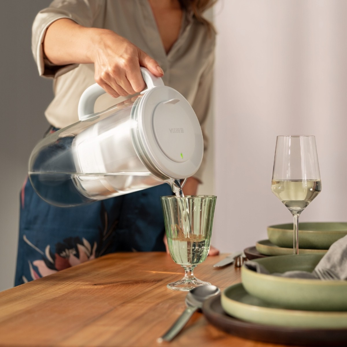 Osoba nalewa wodę z białego dzbanka do zielonej szklanki. Kieliszek do wina i naczynia na drewnianym stole.