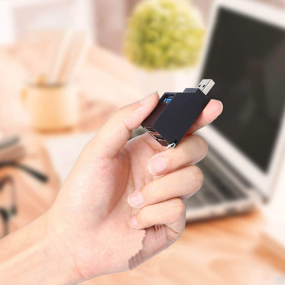 Eine Hand hält einen schwarzen USB-Hub. Ein Laptop und eine verschwommene Pflanze sind im Hintergrund.