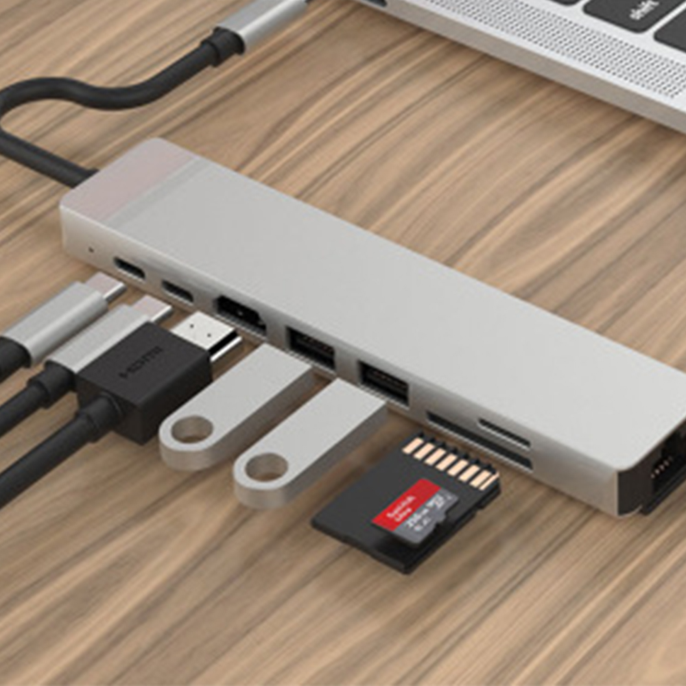 Silberner USB-Hub mit verschiedenen Anschlüssen, verbunden mit einem Laptop. Enthält USB-Laufwerke und eine SD-Karte, auf einer Holzoberfläche.