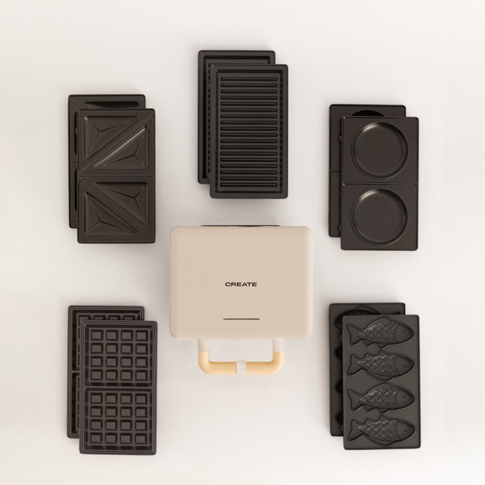 Una sandwichera CREATE beige con varias placas negras para gofres, sándwiches y pasteles con forma de pez sobre un fondo blanco.