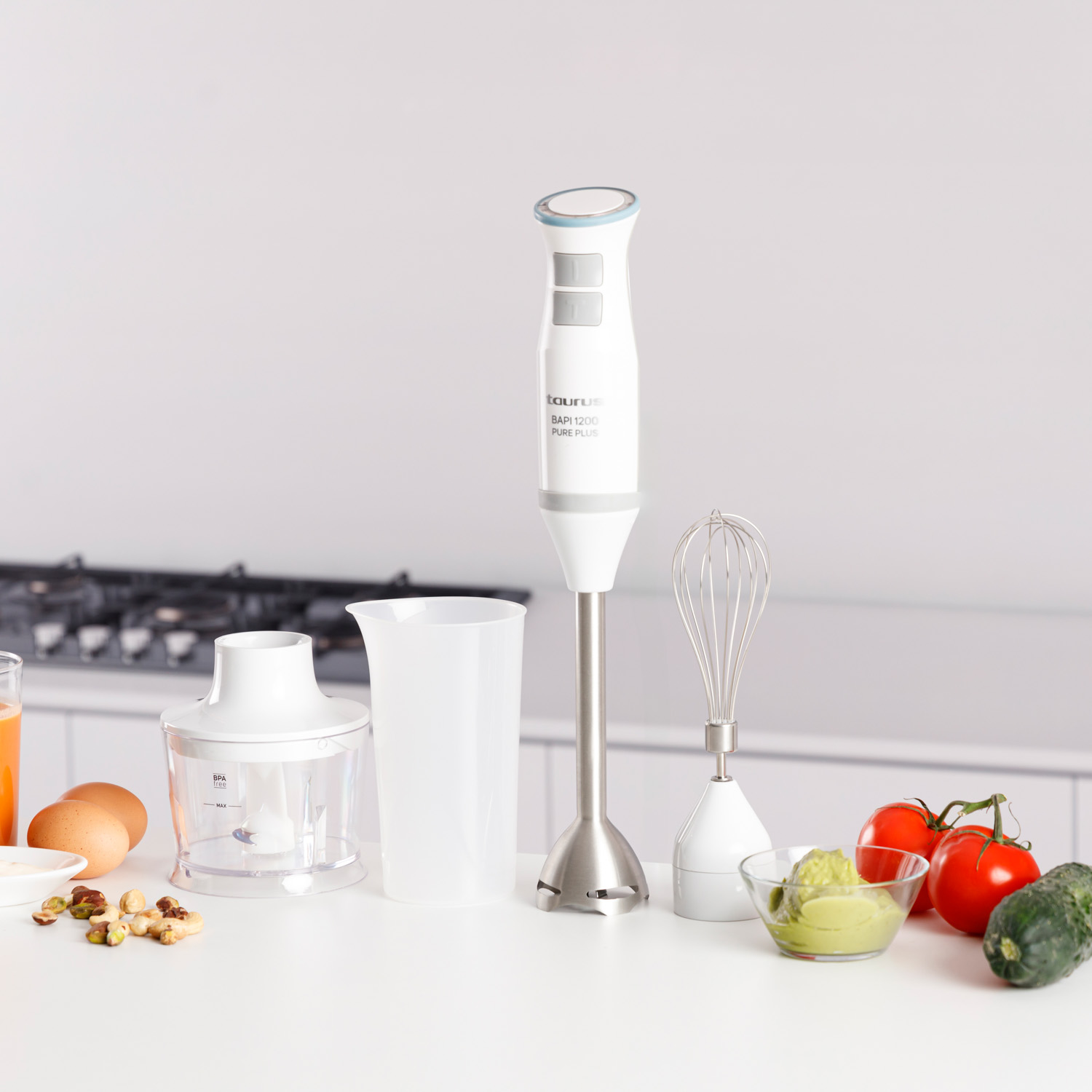 Una batidora blanca con accesorios sobre una superficie blanca con verduras, huevos y nueces. También hay un vaso de zumo.