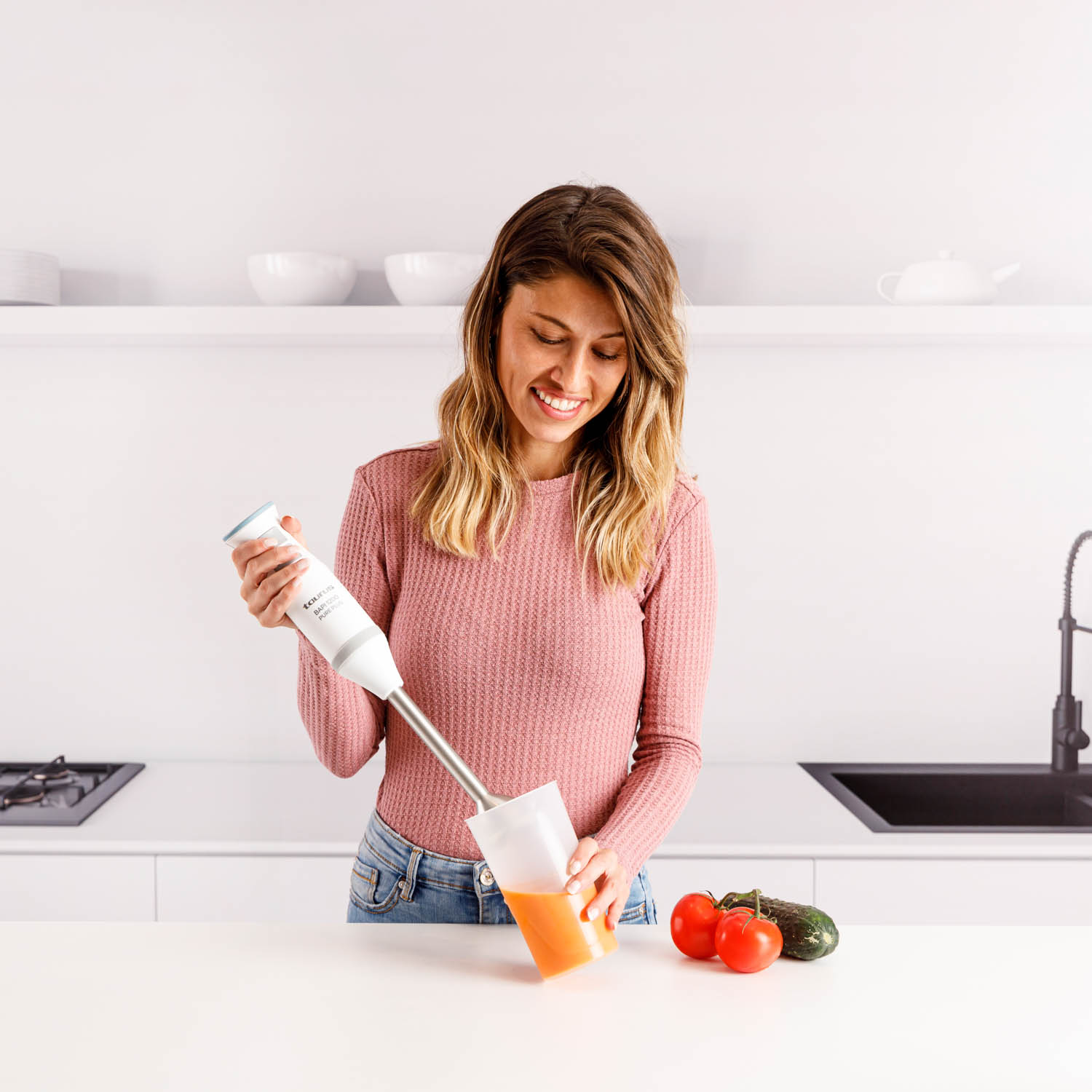 Una mujer sonríe mientras mezcla jugo con una batidora de mano en una cocina. Tomates y un pepino están en el mostrador.