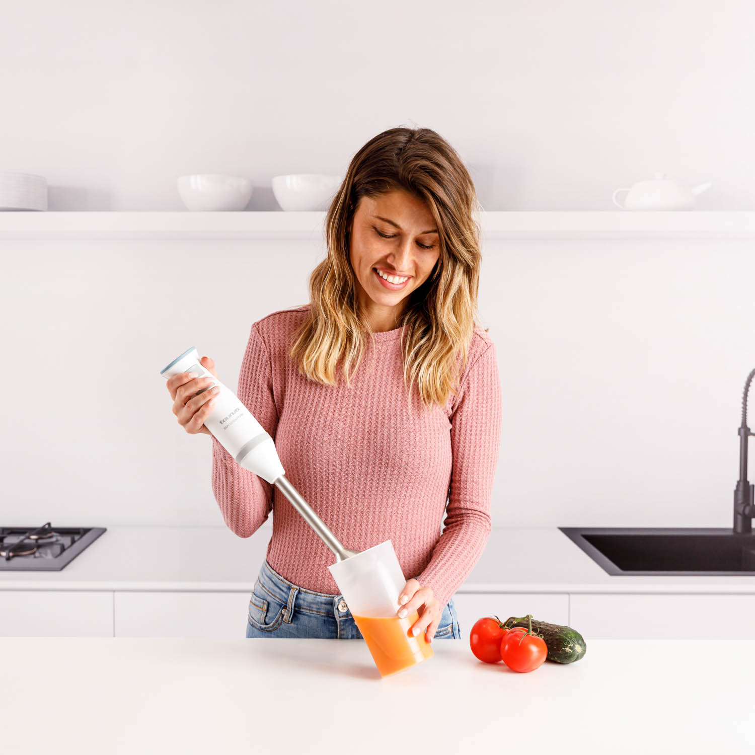 Una mujer sonríe mientras mezcla jugo con una batidora de mano en una cocina. Hay tomates y un pepino en la encimera.