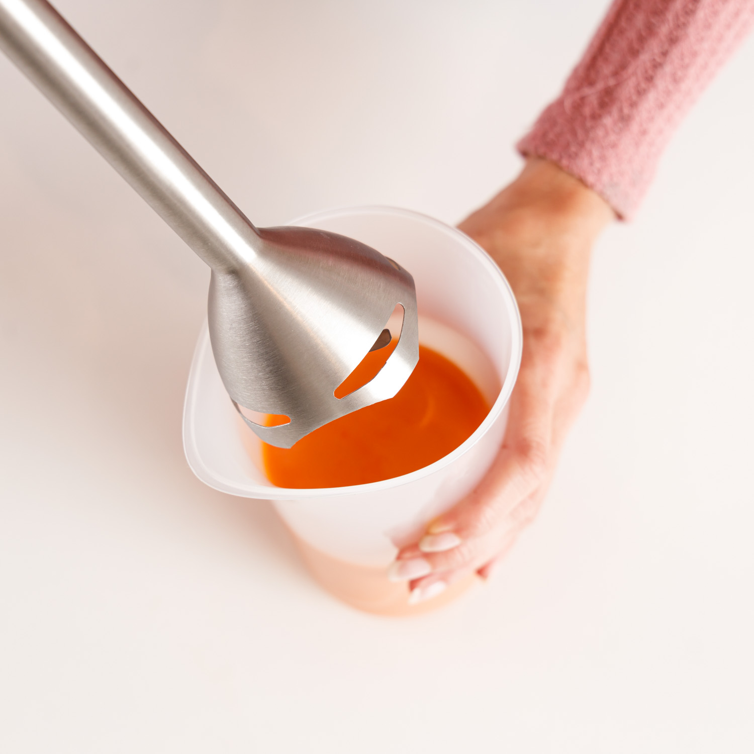 Una mano sostiene una licuadora sobre una taza con líquido naranja. Se ve un suéter rosa. La escena está sobre una superficie blanca.