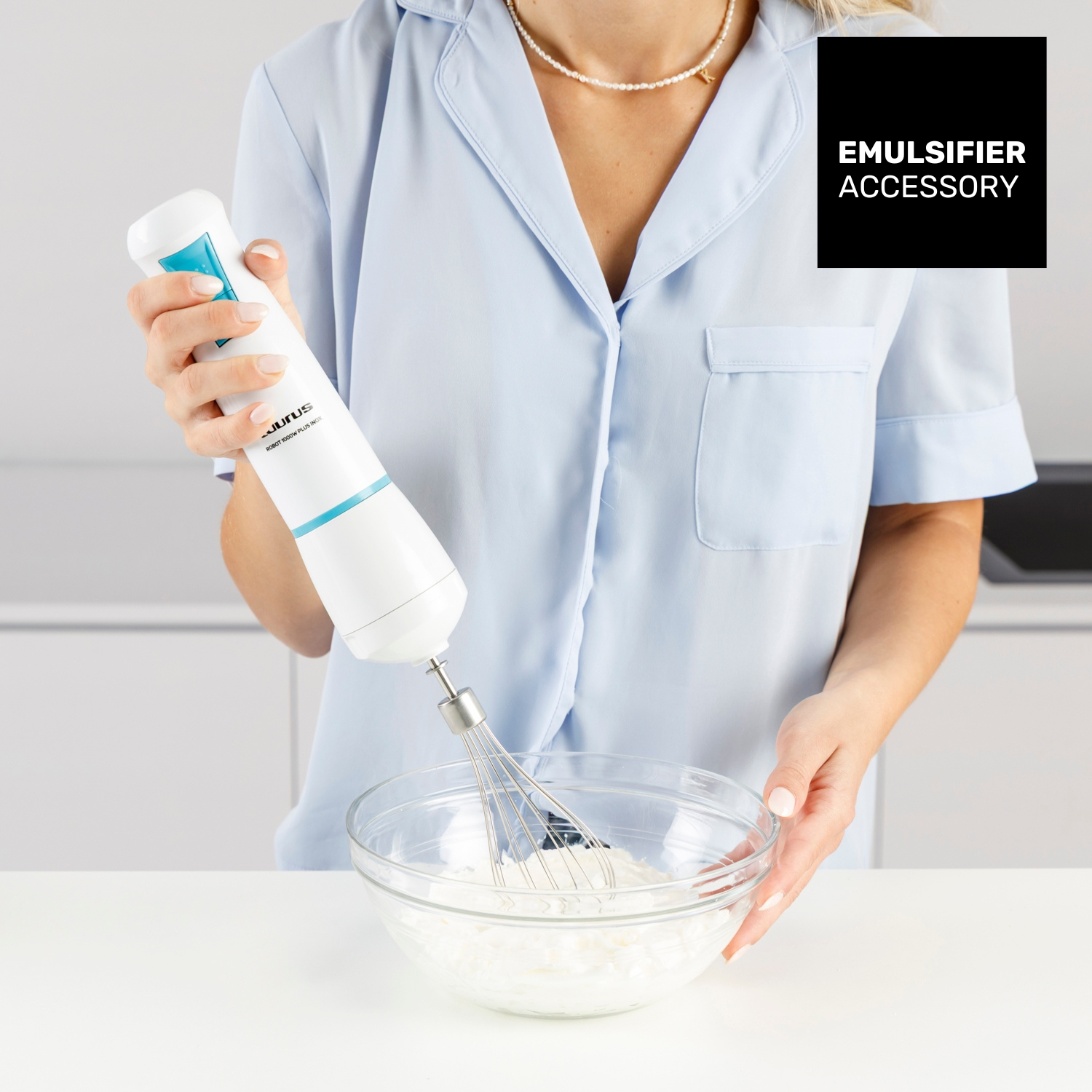 Una persona tiene un frullatore a immersione bianco e blu sopra una ciotola di vetro con una miscela bianca. Lo sfondo mostra una cucina.