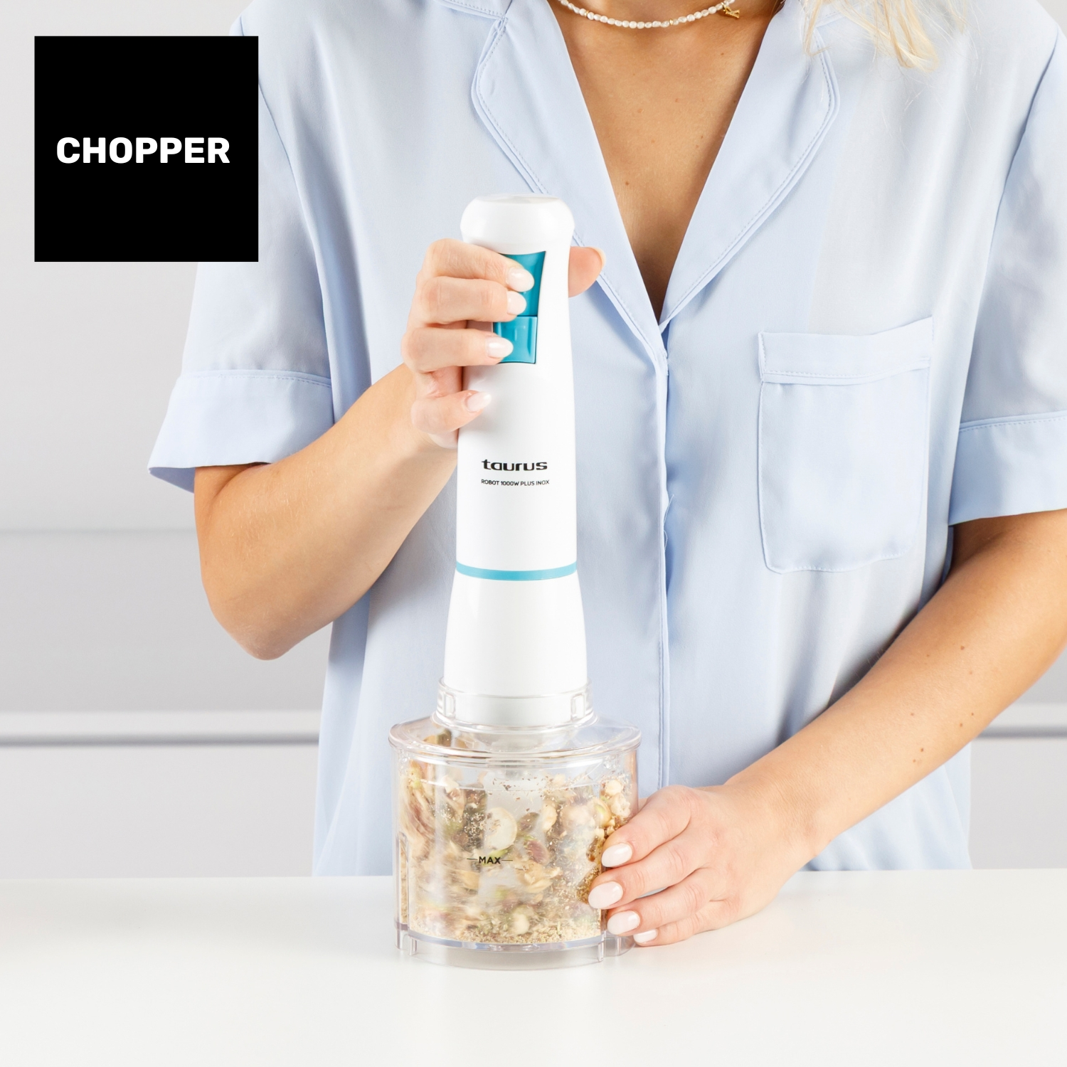 Una donna tiene in mano un tritatutto bianco e blu, che sta tagliando il cibo. Il tritatutto è sopra un contenitore trasparente. La donna indossa una camicia azzurra.