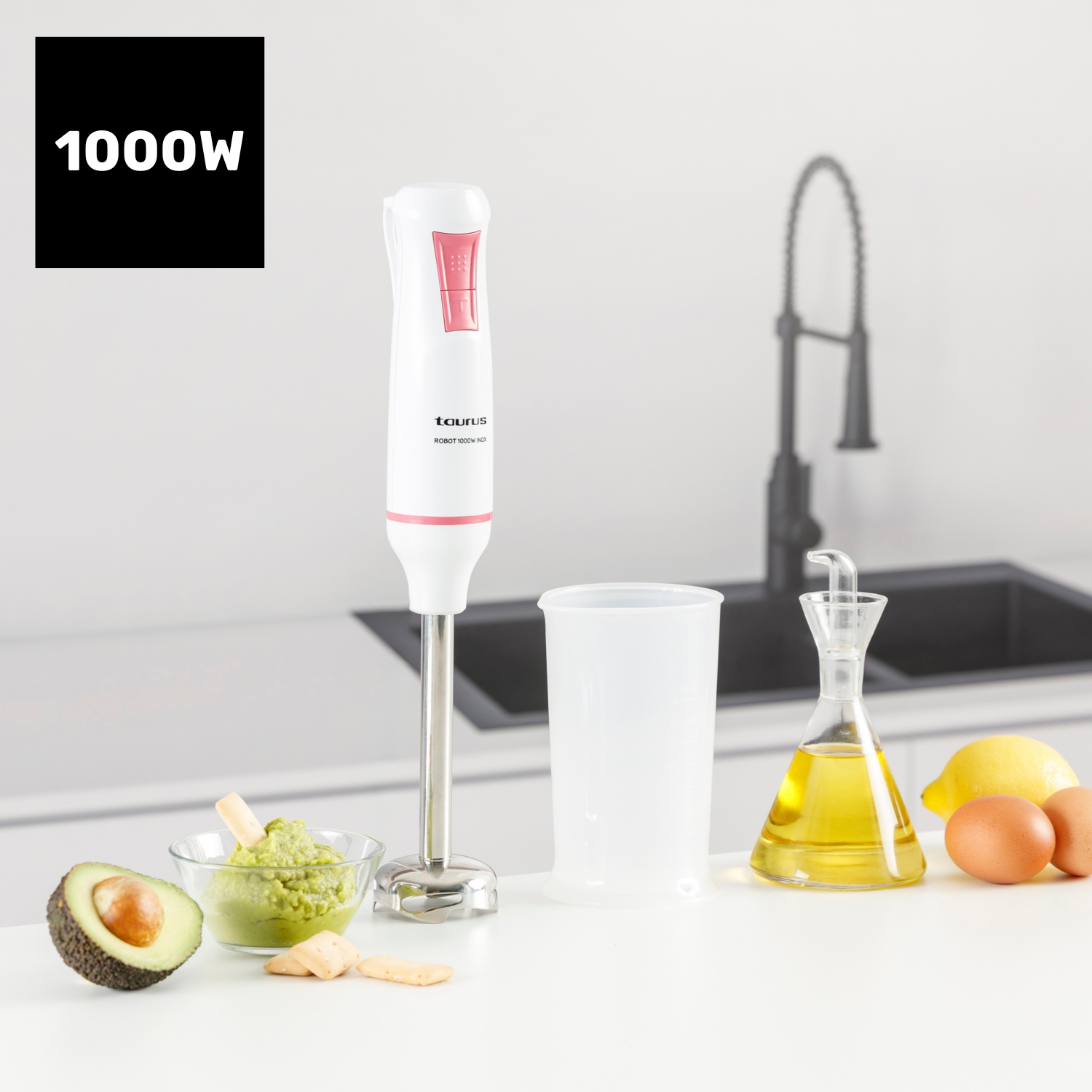 Un frullatore bianco con un accento rosa, avocado, guacamole, olio, limone, uova e una tazza su una superficie bianca.