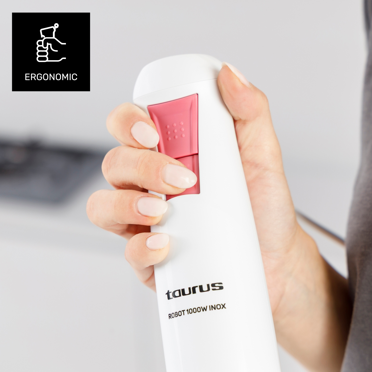 Una mano tiene un frullatore Taurus Robot 1000W Inox bianco. Ha un pulsante rosa. La parola "Ergonomic" è scritta a sinistra.