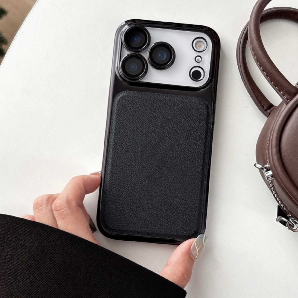 Una mano tiene una custodia nera per telefono con texture in pelle. Una borsa marrone sullo sfondo su una superficie bianca.