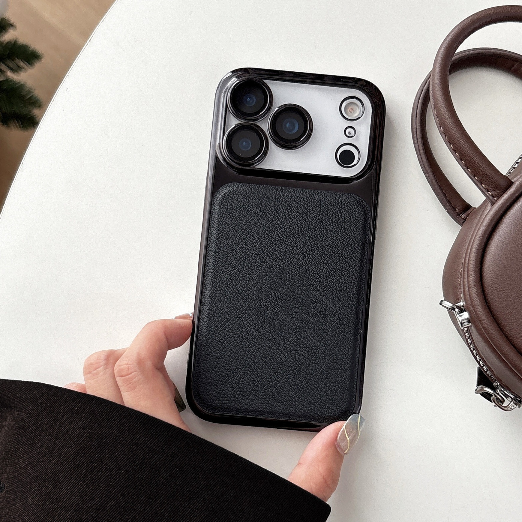 Una mano che tiene una custodia nera per telefono con texture in pelle, accanto a una borsa marrone su una superficie bianca.
