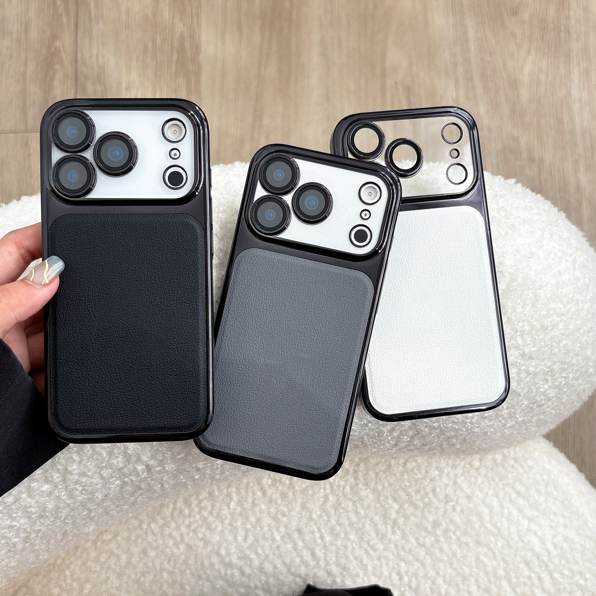 Tre custodie per telefono: nera, grigia e bianca. Ognuna ha un modulo fotocamera e un retro simile alla pelle, su una superficie bianca e soffice.