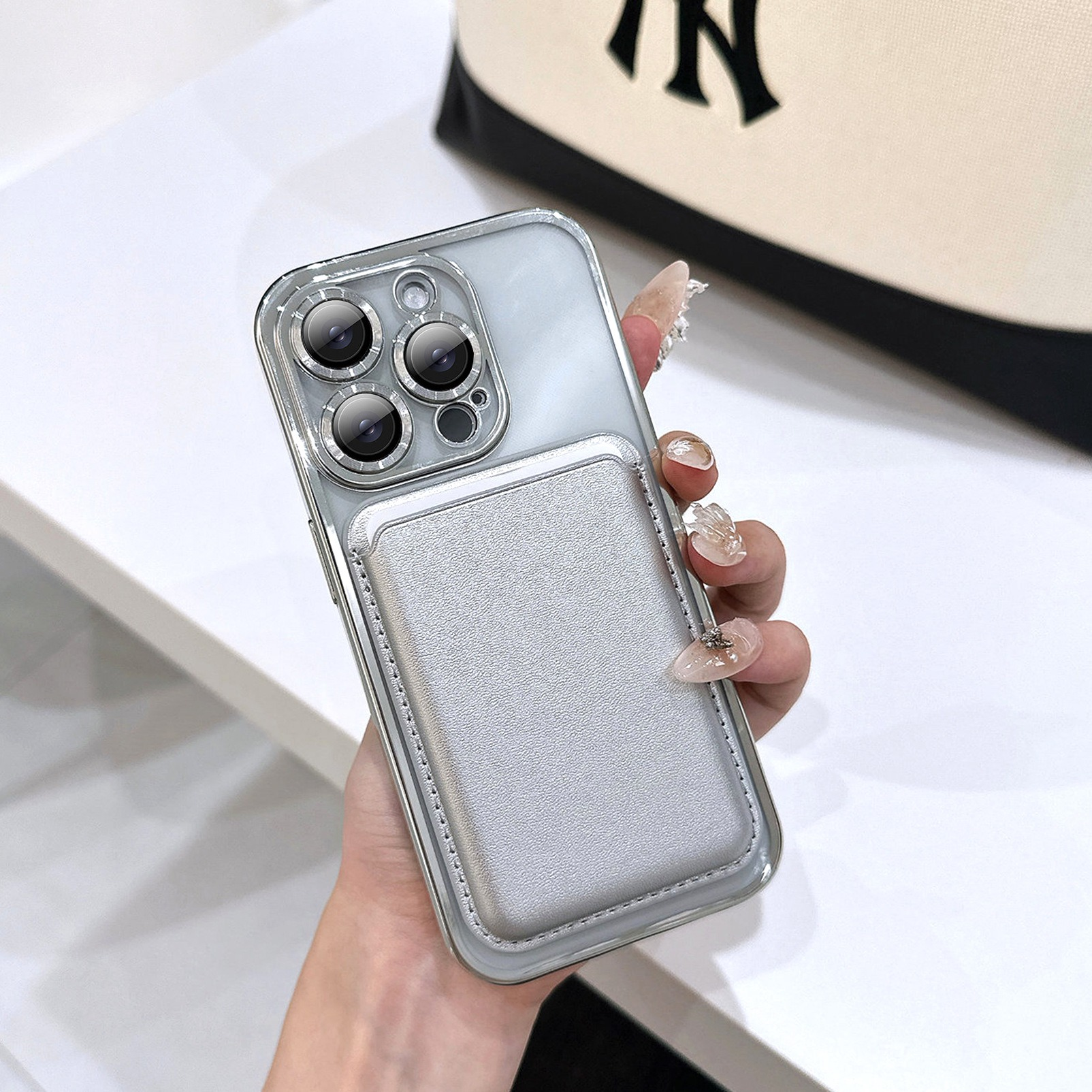 Una mano tiene un telefono argento con un porta carte. Le unghie sono curate. Una borsa con un logo è sullo sfondo.