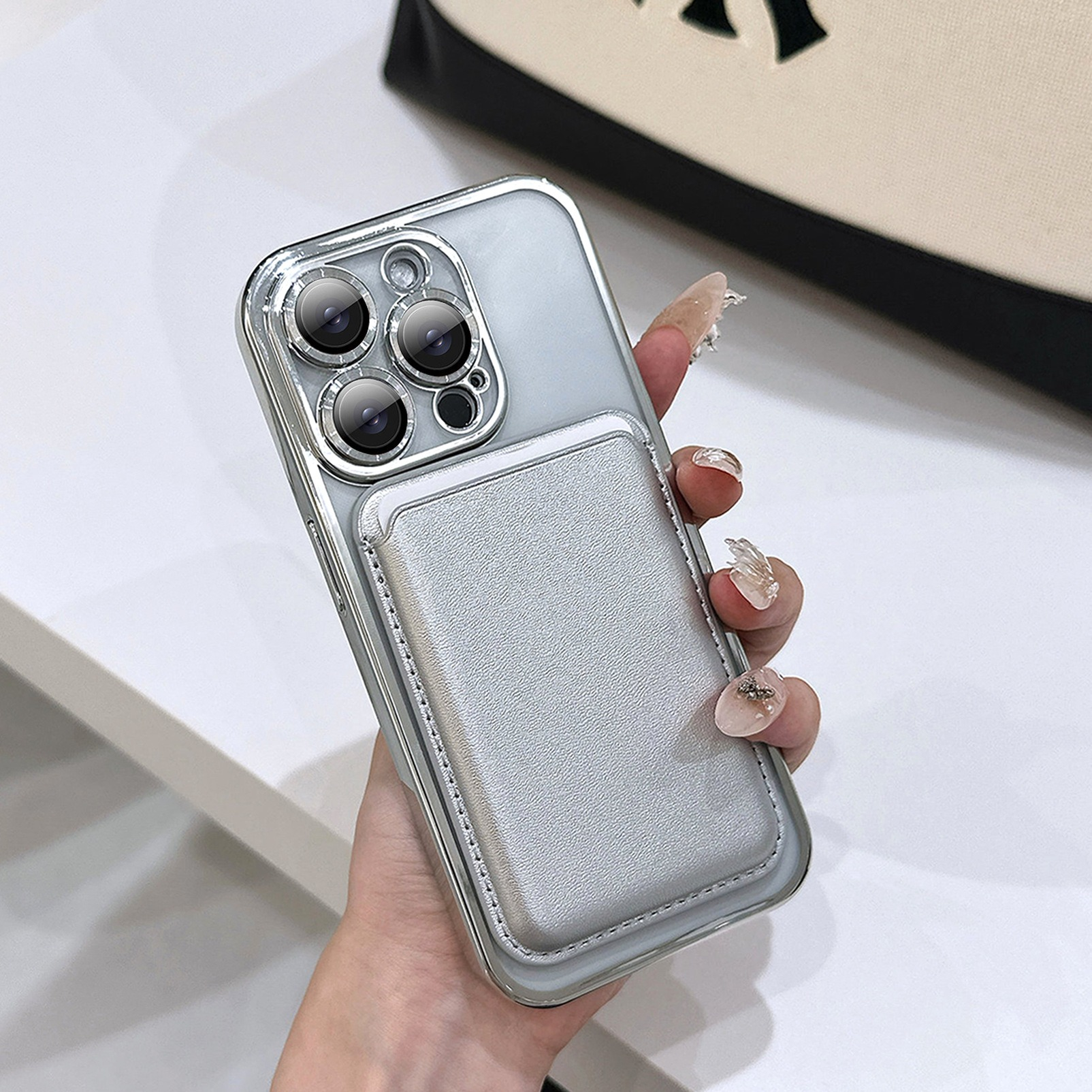 Una mano tiene un telefono argento con un porta carte. La custodia del telefono ha tre lenti. Lo sfondo è sfocato.