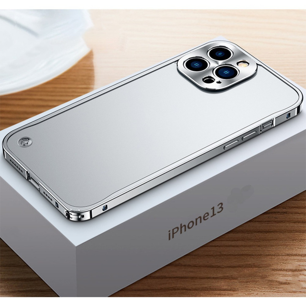 Eine silberne iPhone 13-Hülle liegt auf ihrer weißen Schachtel, die Kameralinsen des Telefons sind sichtbar. Auf der Schachtel steht 'iPhone13'.