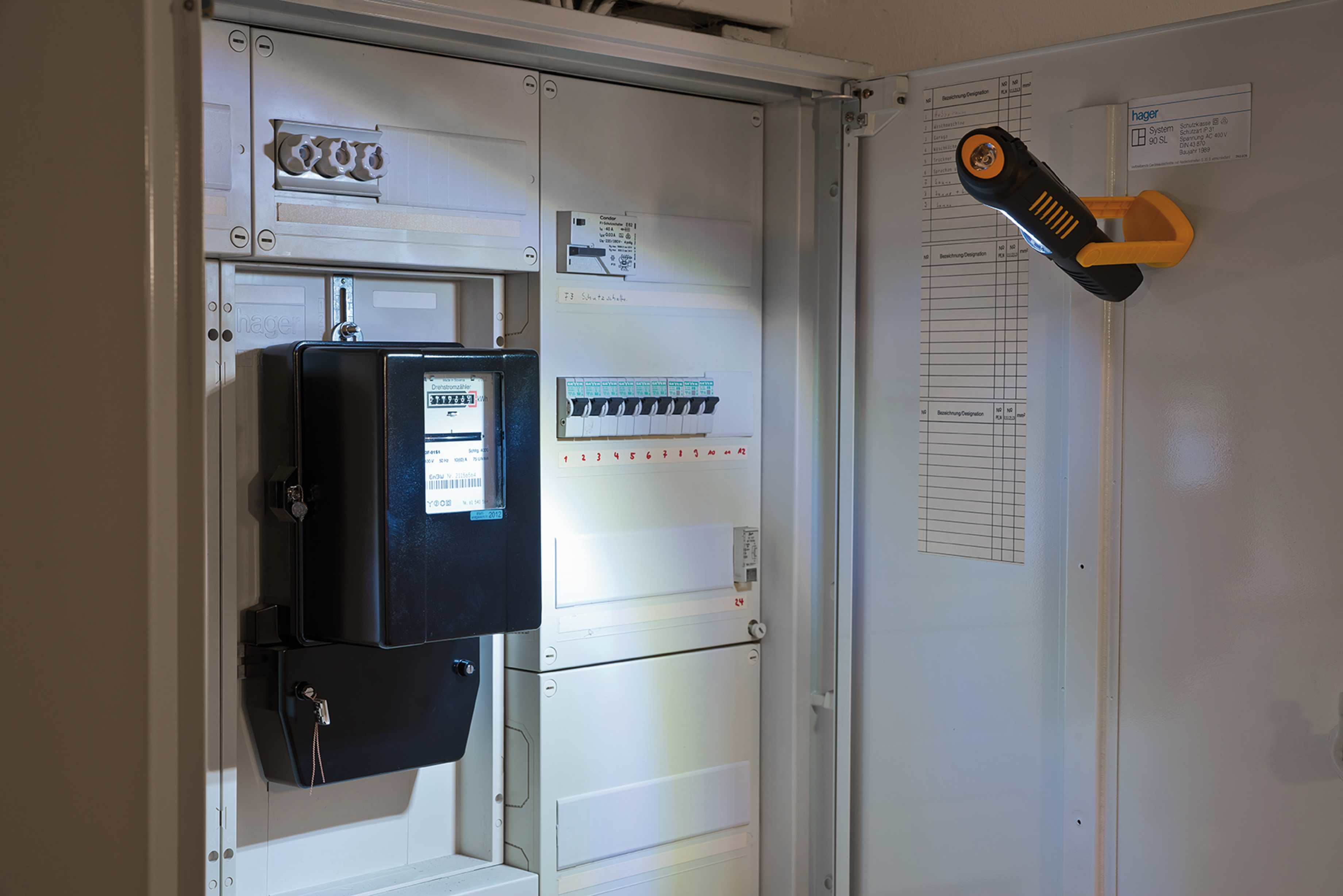 Contatore elettrico e quadro elettrico. Una torcia è appesa alla porta. Numeri ed etichette visibili.