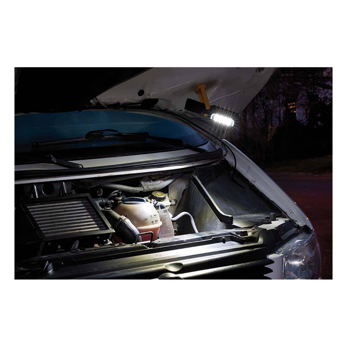 Un vano motore di un'auto è aperto, illuminato da una luce da lavoro. Il cofano è sollevato. Lo sfondo è scuro con alberi.