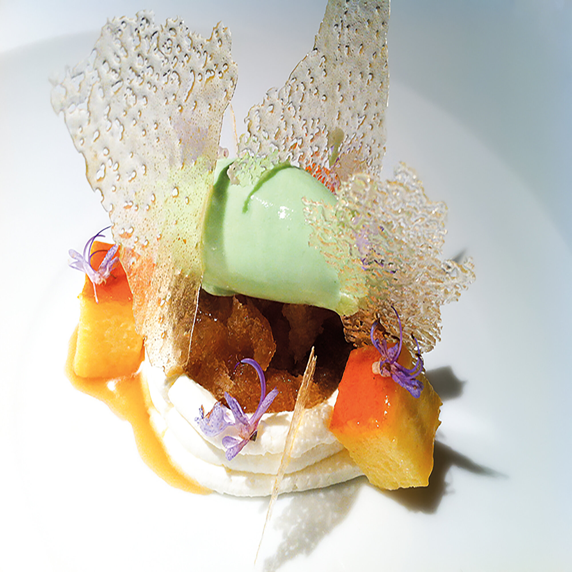 Postre en plato blanco. Bola de helado verde, trozos de azúcar, crema blanca, cubos naranjas y una guarnición transparente.