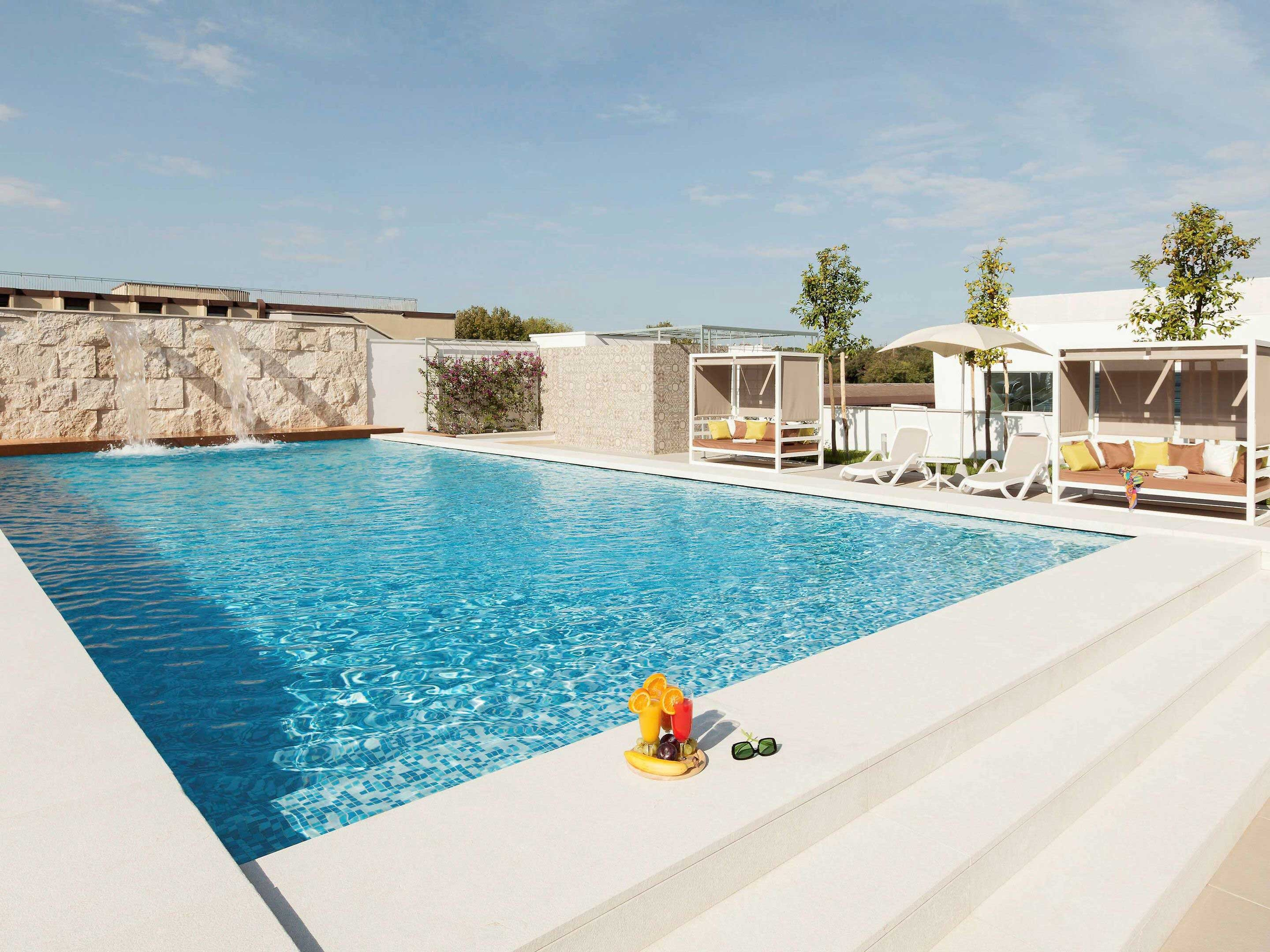 Escena junto a la piscina con agua azul, escalones blancos y una bandeja con bebidas y frutas. Se ven cenadores y árboles.