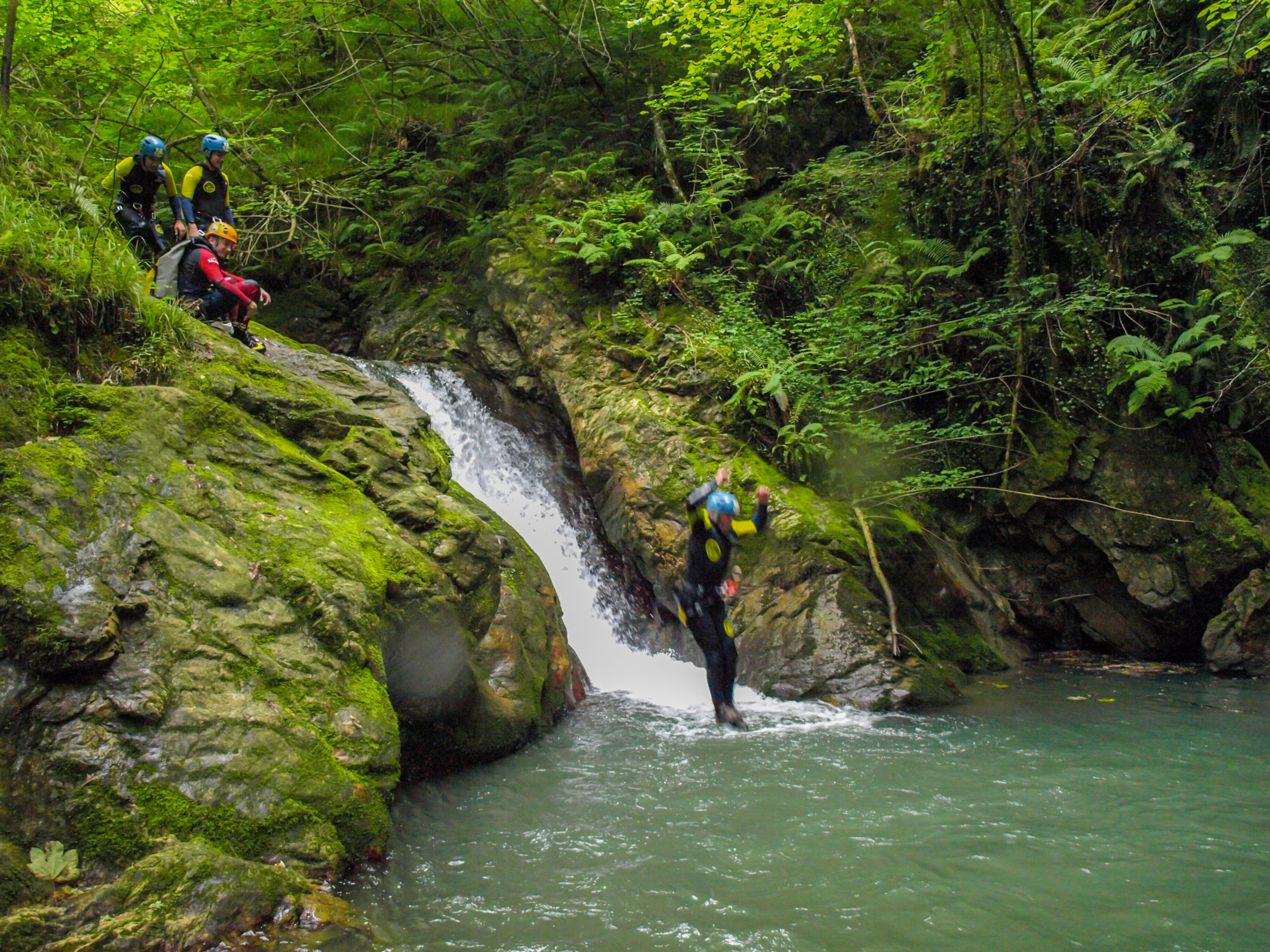 Una persona salta al agua mientras otros observan. El bosque verde rodea la cascada y el agua.