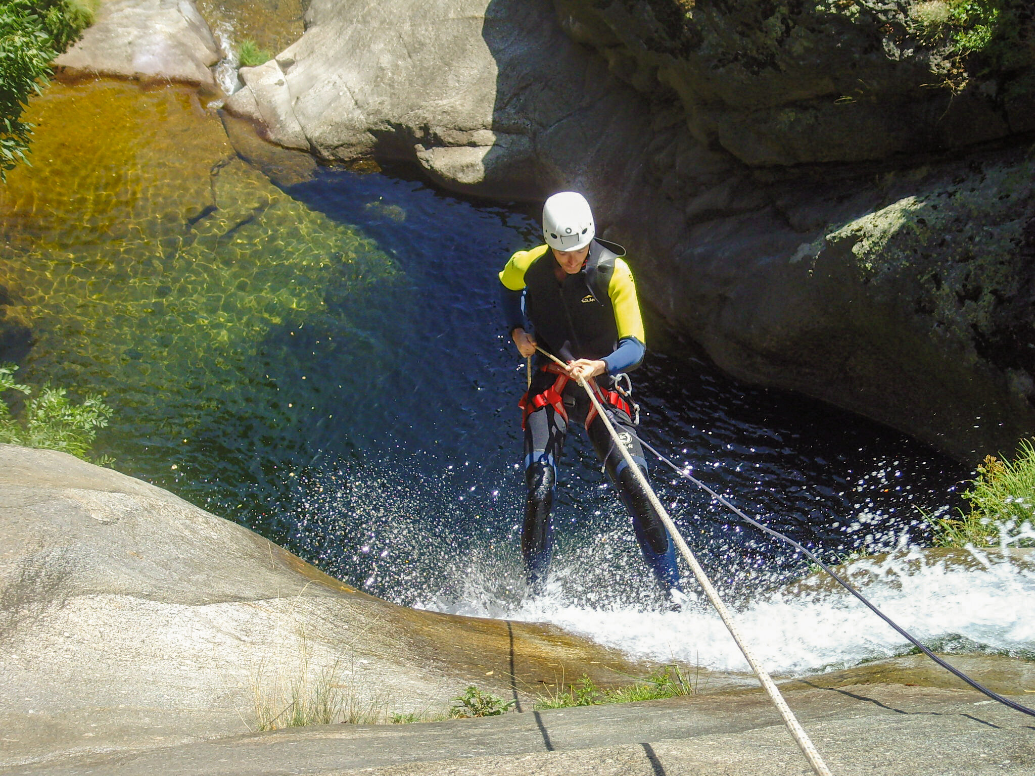 Una persona desciende en rápel por una cascada. Lleva casco y traje de neopreno. Agua clara y rocas la rodean.