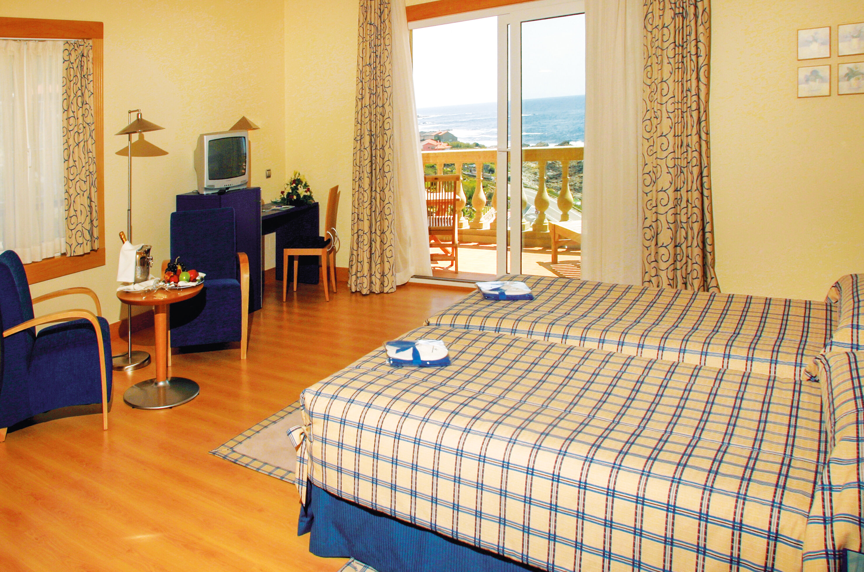 Una habitación de hotel con dos camas, balcón con vista al mar, mesa, sillas y TV.