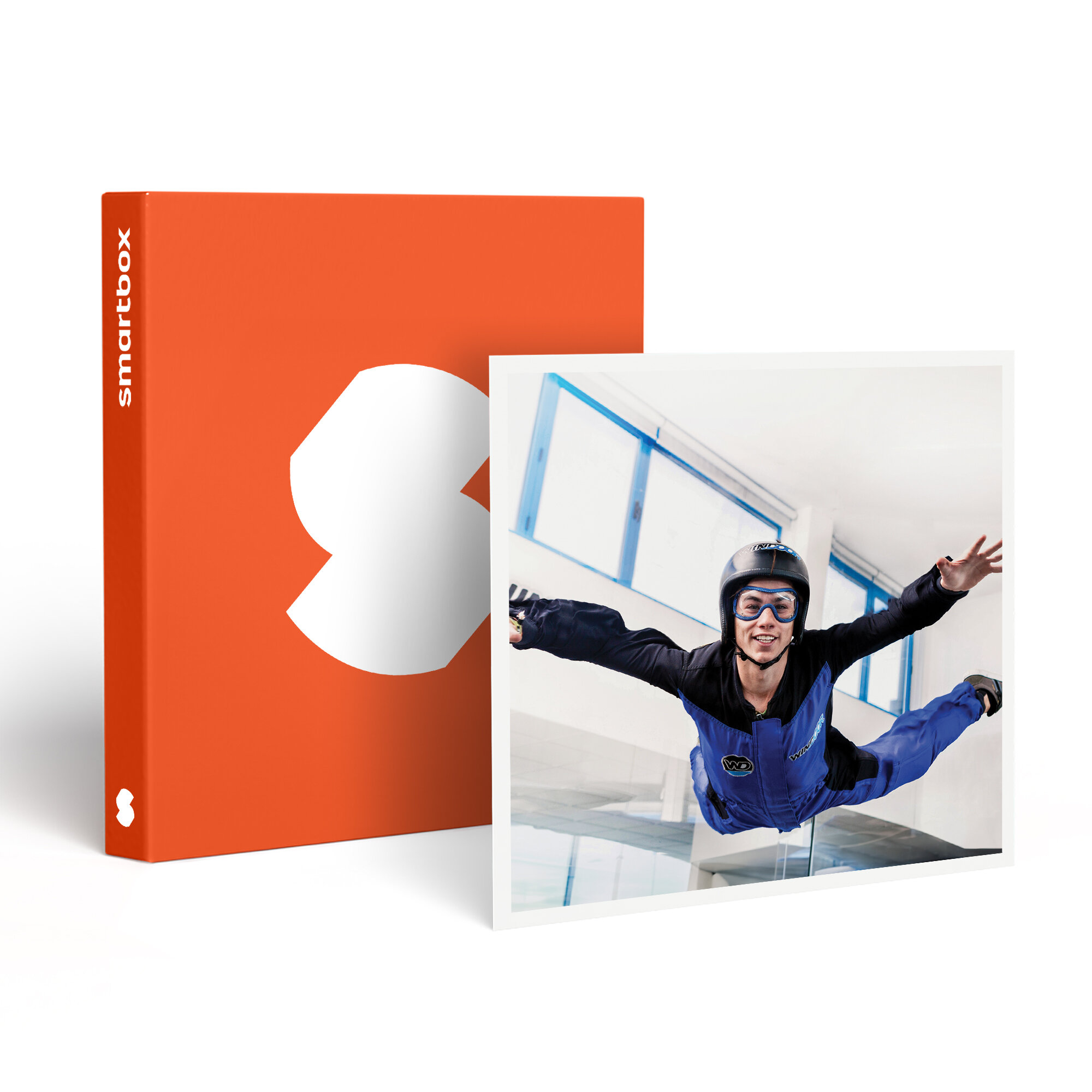 Hombre con mono azul y casco, volando en un túnel de viento. Caja naranja con logo blanco a la izquierda.