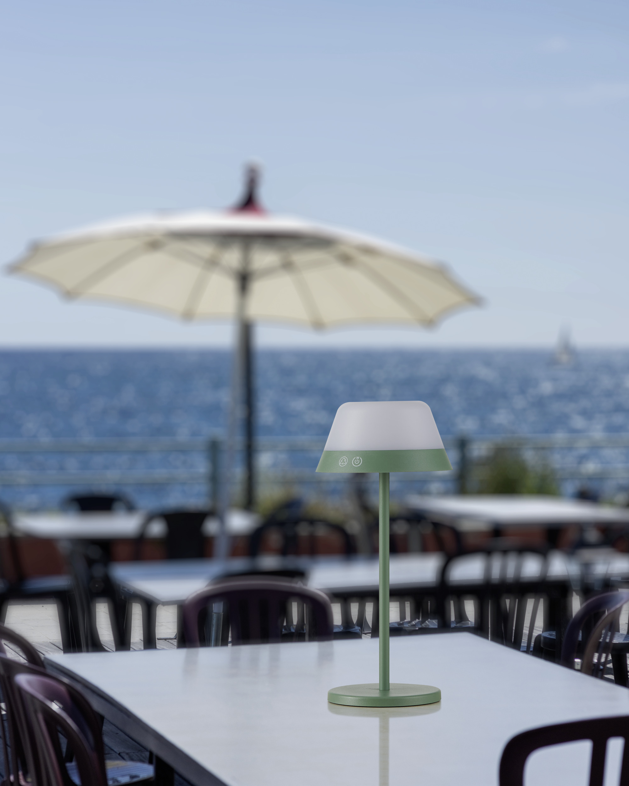 Eine grün-weiße Lampe steht auf einem weißen Tisch. Im Hintergrund sind Tische, Stühle und das Meer.