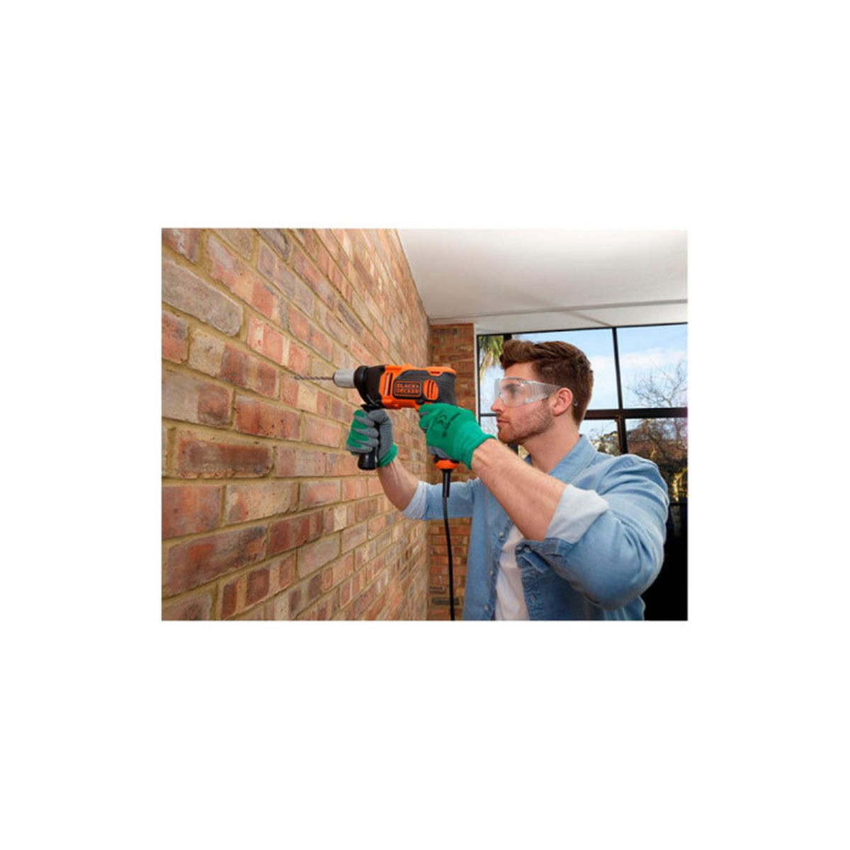 Mężczyzna wierci w ceglanym murze pomarańczowo-czarną wiertarką. Nosi okulary ochronne i zielone rękawice.
