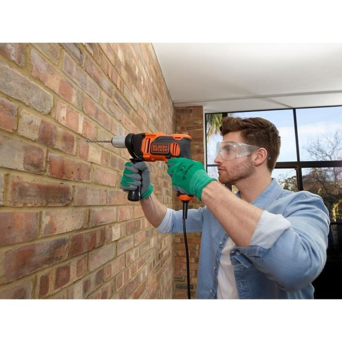 Mężczyzna wierci w ceglanym murze wiertarką Black+Decker, nosząc okulary ochronne i zielone rękawice.