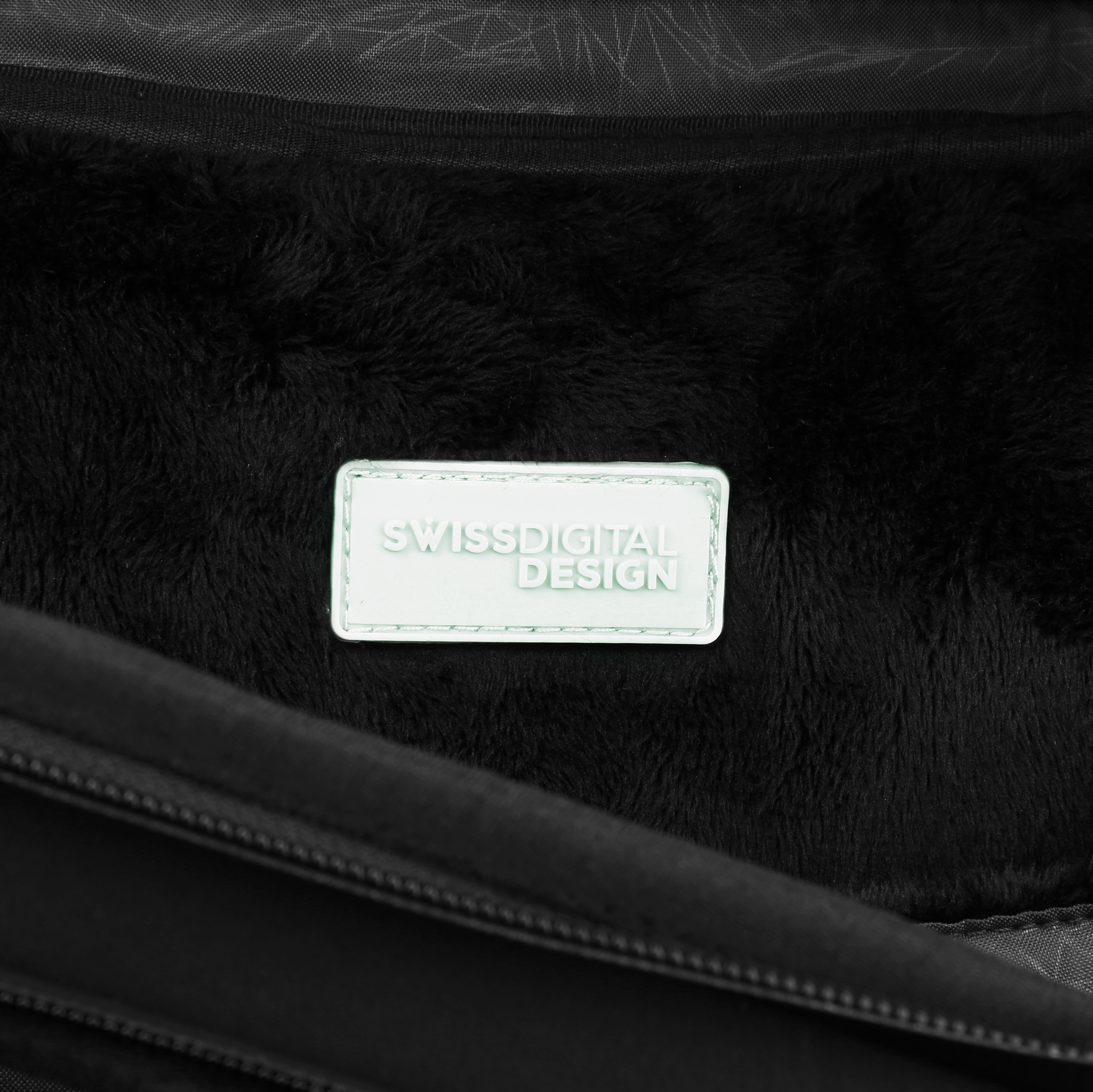 Primo piano di un interno nero e soffice. Un'etichetta verde chiaro recita SWISSDIGITAL DESIGN. La cerniera di una borsa è visibile.