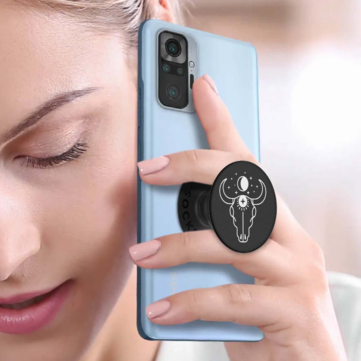 Eine Frau hält ein hellblaues Telefon mit einem schwarzen Popsocket mit einem Bullenschädel-Design. Ihre Nägel sind rosa lackiert.