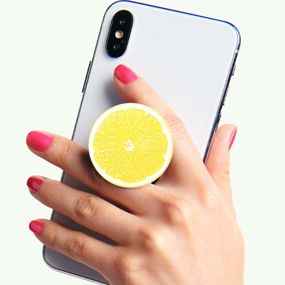 Una mano sostiene un smartphone con un agarre de teléfono de rodaja de limón. También son visibles uñas rosas y una funda blanca para el teléfono.