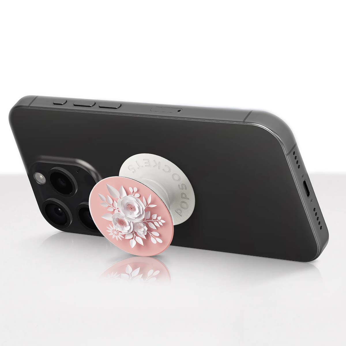 Smartphone nero con PopSocket rosa con design floreale bianco, appoggiato su una superficie riflettente.