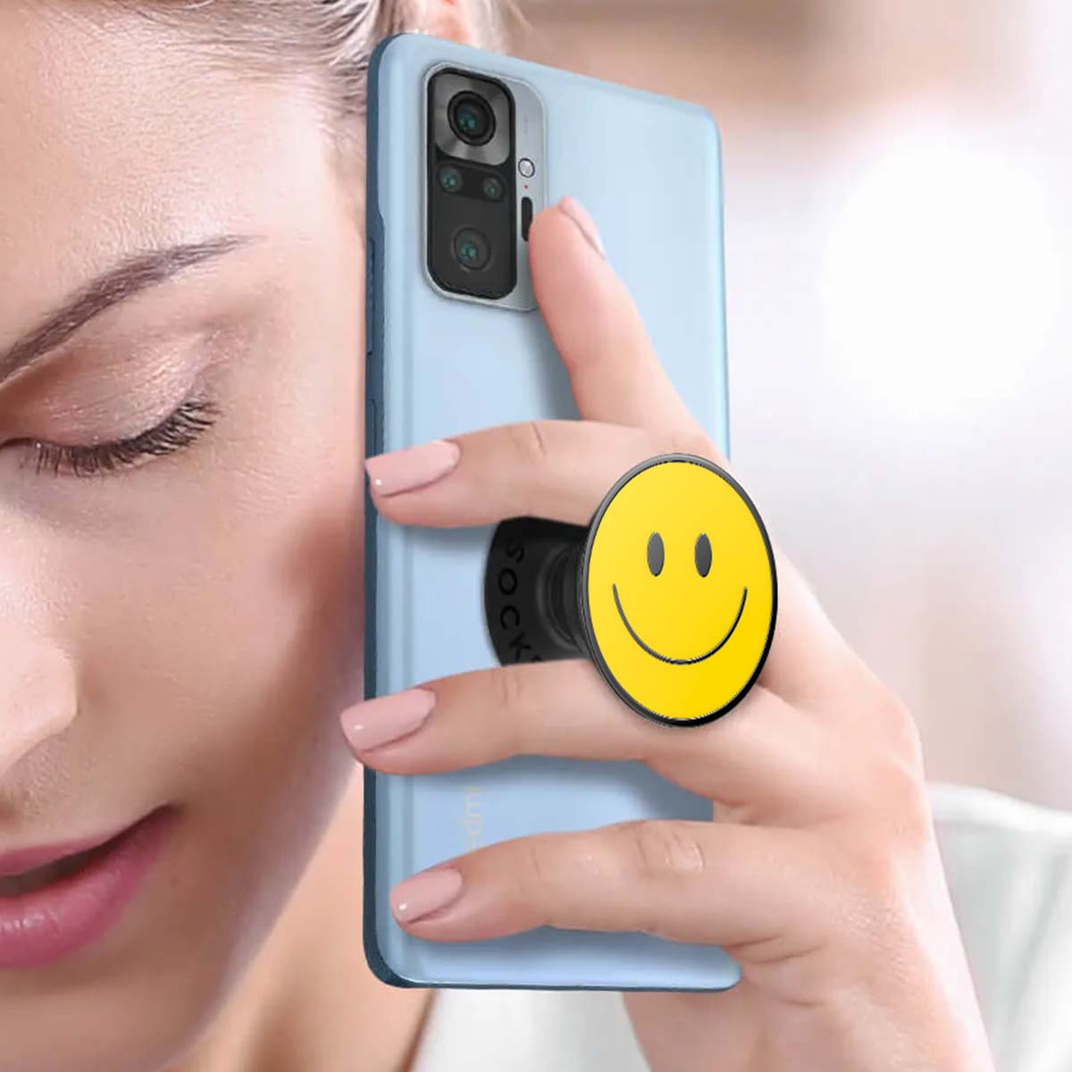 Una donna tiene in mano un telefono azzurro con un pop socket a forma di faccina sorridente. Le sue unghie sono dipinte di rosa.