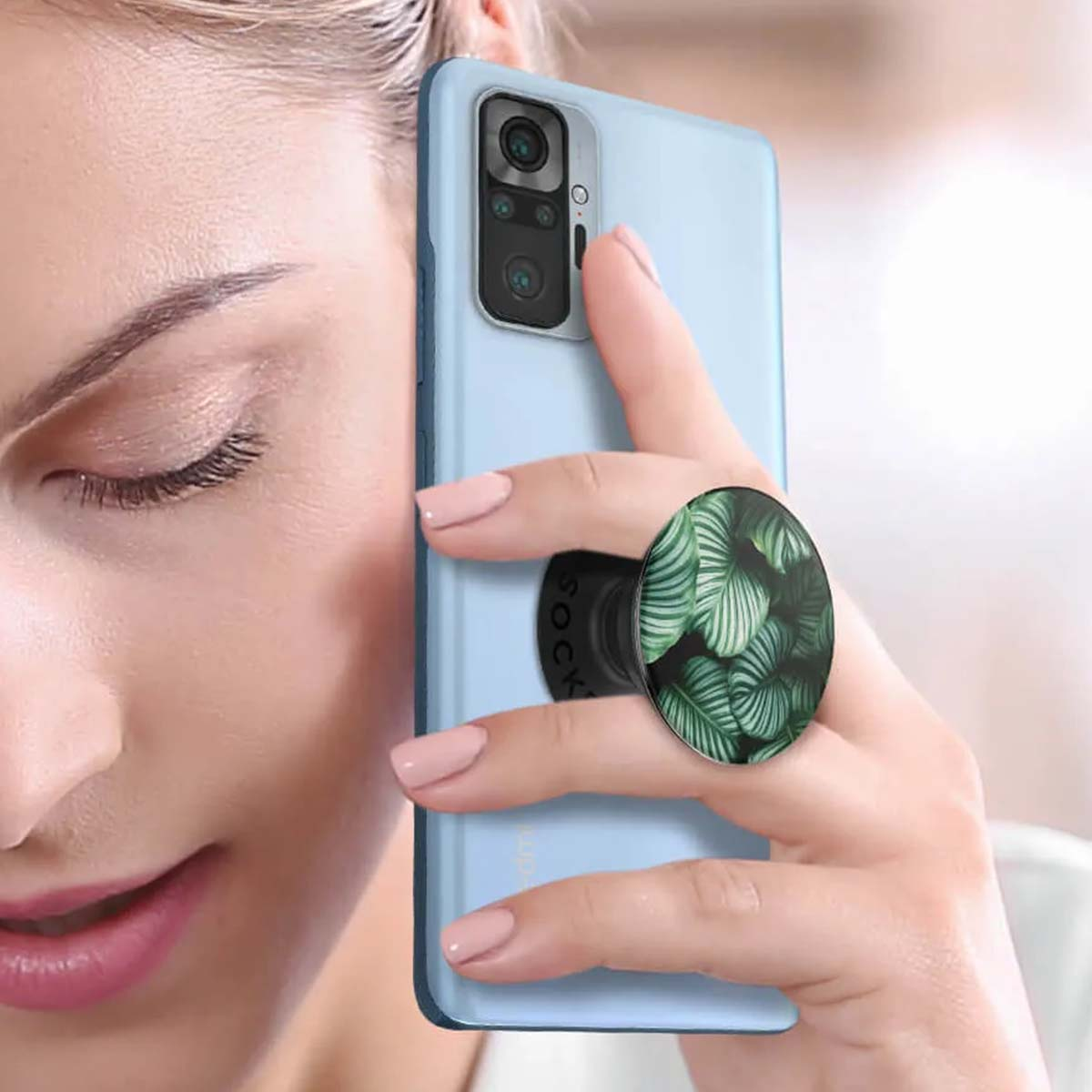 Eine Frau hält ein hellblaues Telefon mit einem Pop-Socket mit grünem Blattdesign. Ihre Nägel sind rosa lackiert.