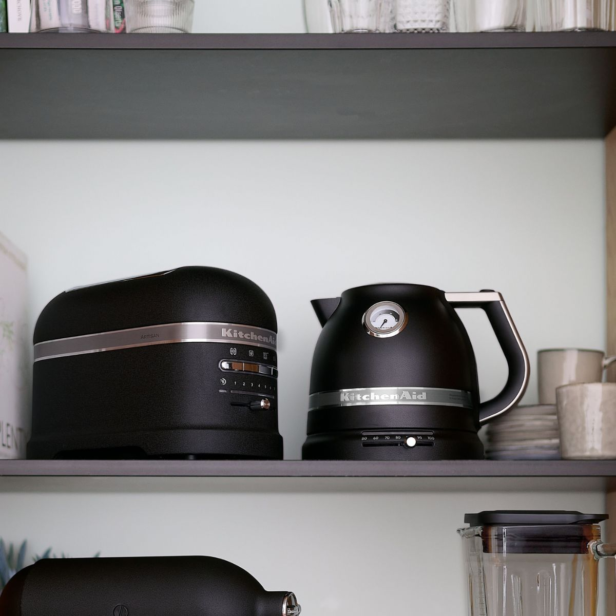 Zwei schwarze KitchenAid-Geräte stehen auf einem Regal. Ein Toaster und ein Wasserkocher mit Temperaturanzeige. Im Hintergrund sind andere Küchenartikel zu sehen.