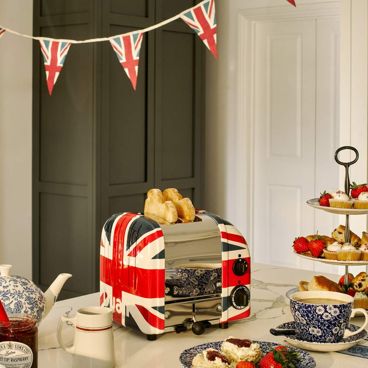 Ein Toaster mit Union-Jack-Design steht auf einer Theke mit Brot. Teeservice, Scones, Erdbeeren und Wimpel vervollständigen die Szene.