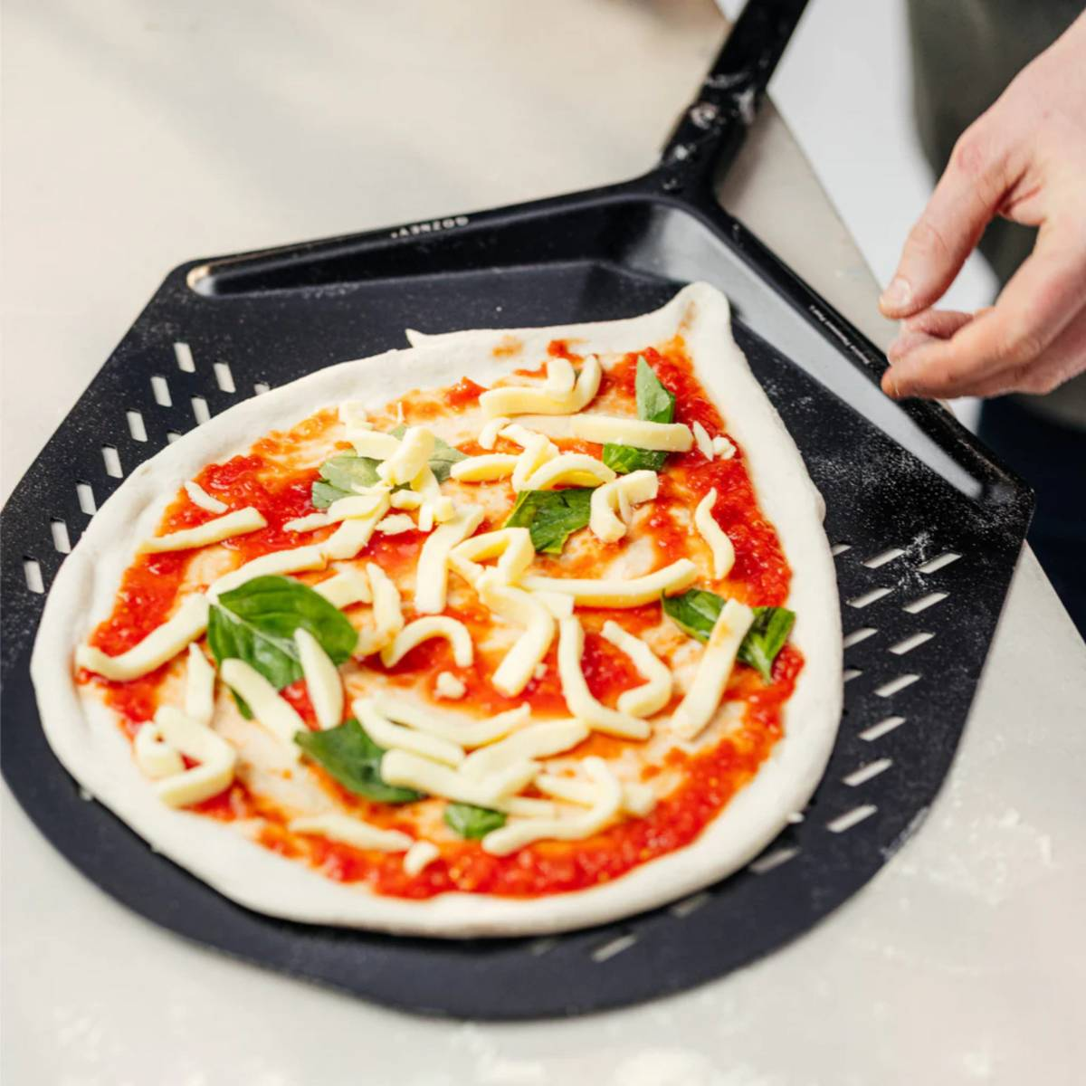 Pizza auf einem Pizzaschieber. Tomatensauce, Käse und Basilikum auf dem Teig. Eine Hand hält den Schieber.