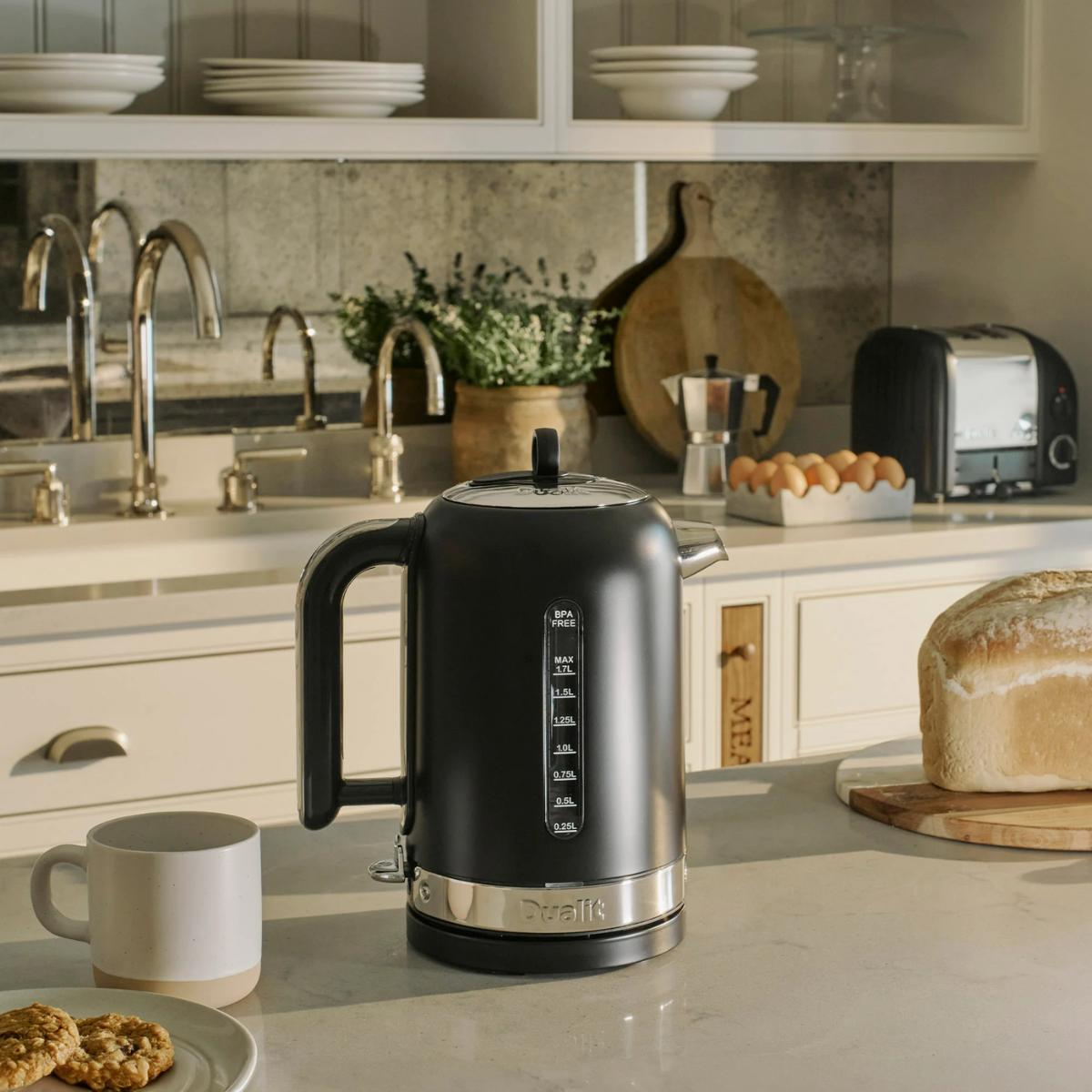 Eine Küchenszene mit einem schwarzen Wasserkocher, Brot und einer Tasse. Im Hintergrund befinden sich ein Toaster, Eier und ein Schneidebrett aus Holz.