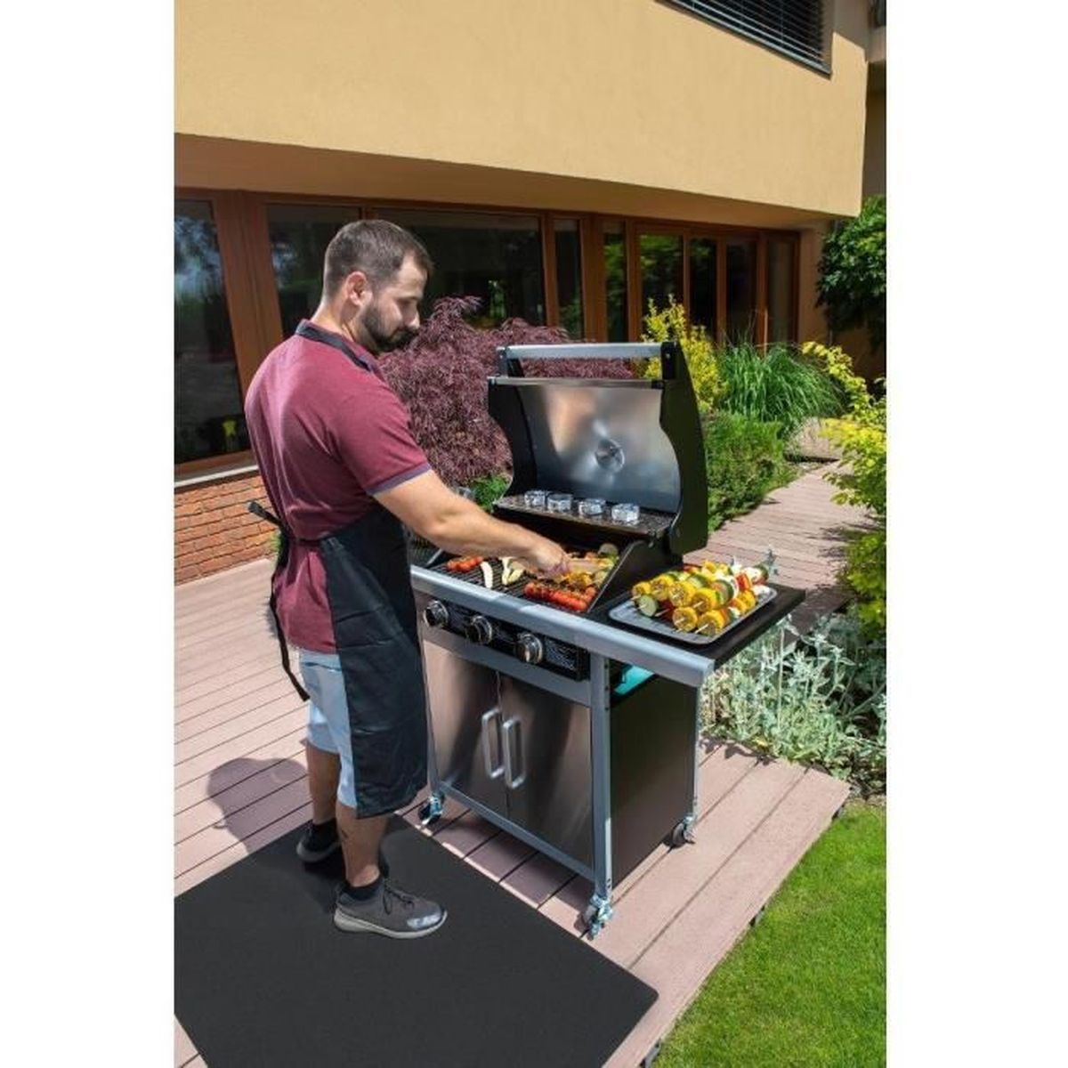 Mann grillt Gemüse auf einem Grill. Er trägt ein rotes Hemd, eine schwarze Schürze und steht auf einer schwarzen Matte. Haus und Garten im Hintergrund.
