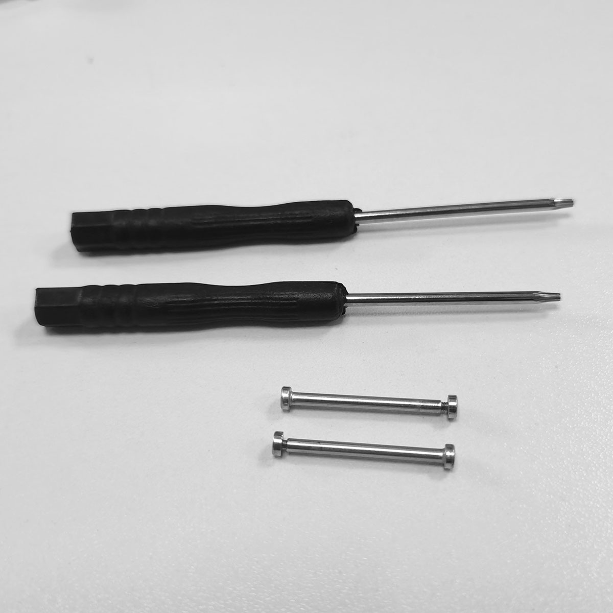 Zwei Schraubendreher mit schwarzen Griffen und silbernen Schäften sowie zwei silberne Metallstäbe auf einer weißen Oberfläche.