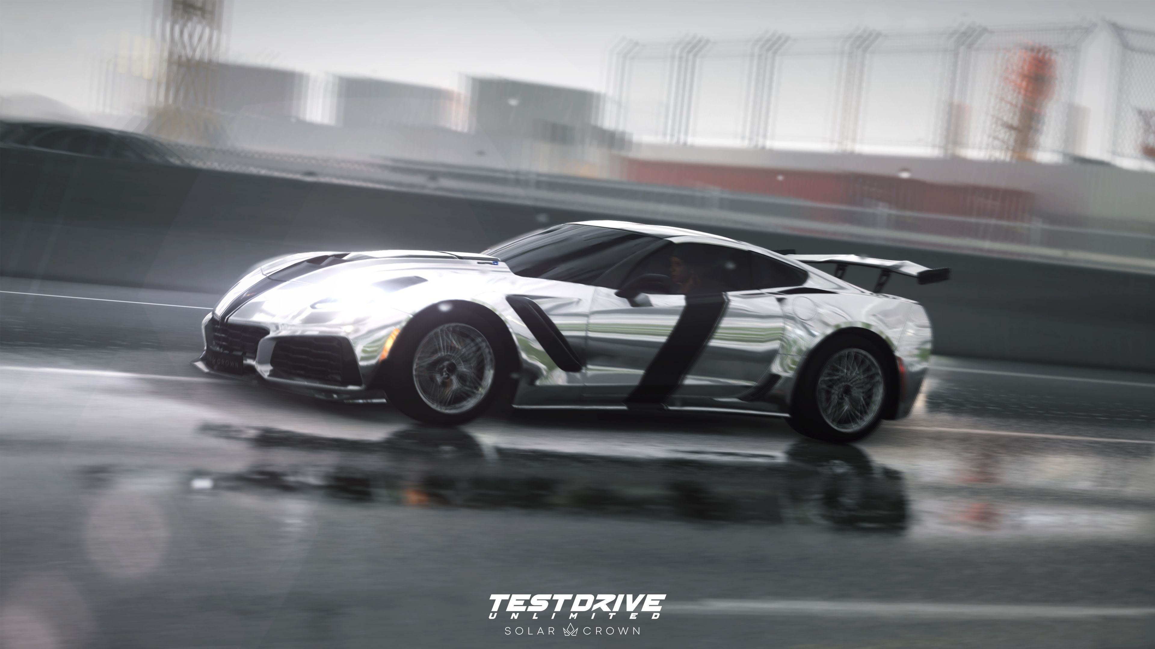 Un'auto sportiva argentata con strisce nere corre su pista bagnata. Sfondo sfocato con edifici e una recinzione.