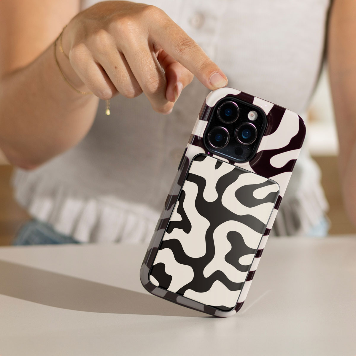 Een hand raakt een telefoon aan met een zwart-witte koeienprint hoes. De telefoon heeft een camerabult. De achtergrond is wazig.
