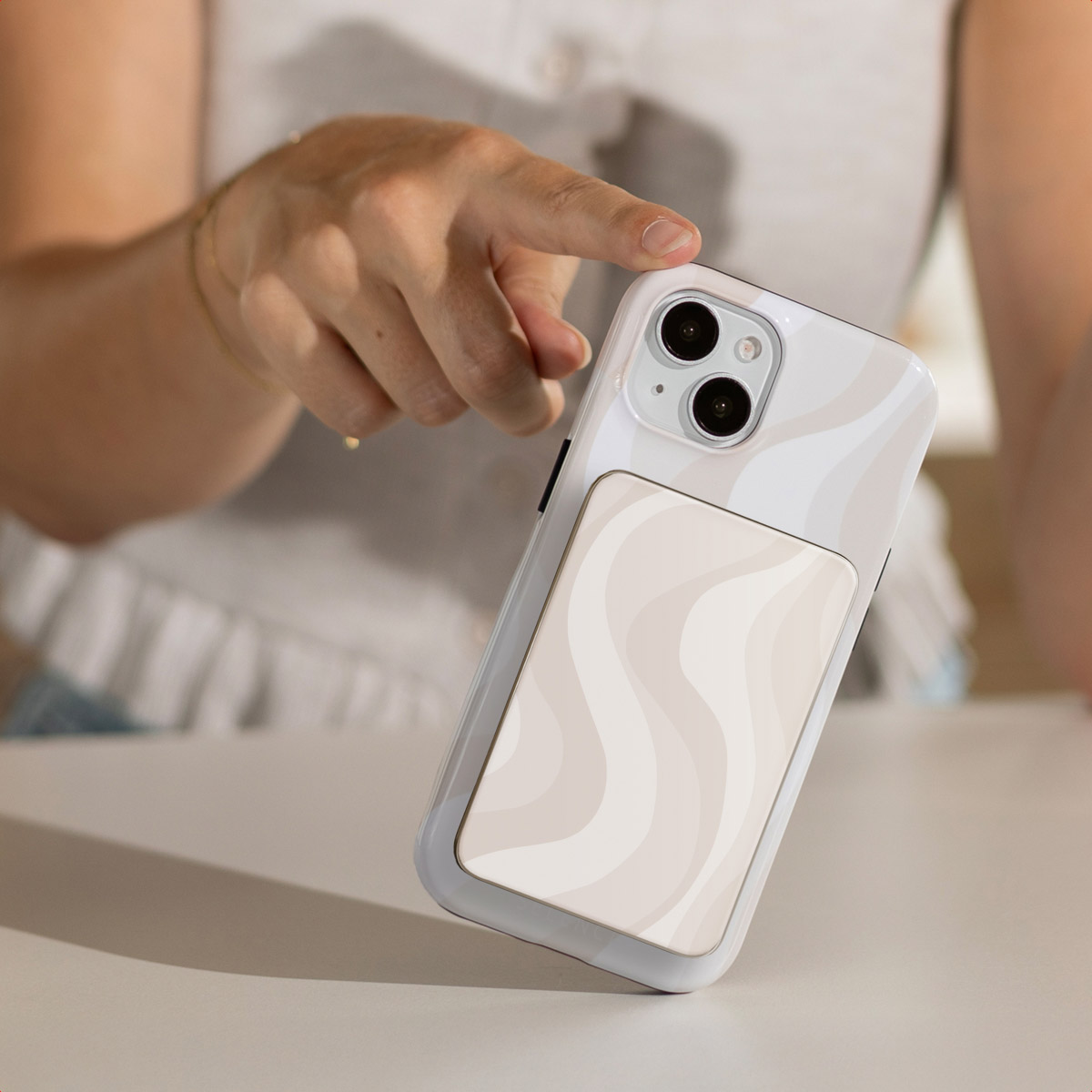Een hand raakt een witte telefoonhoes met een beige golfontwerp aan. De telefoon ligt op een tafel. Persoon draagt een wit overhemd.