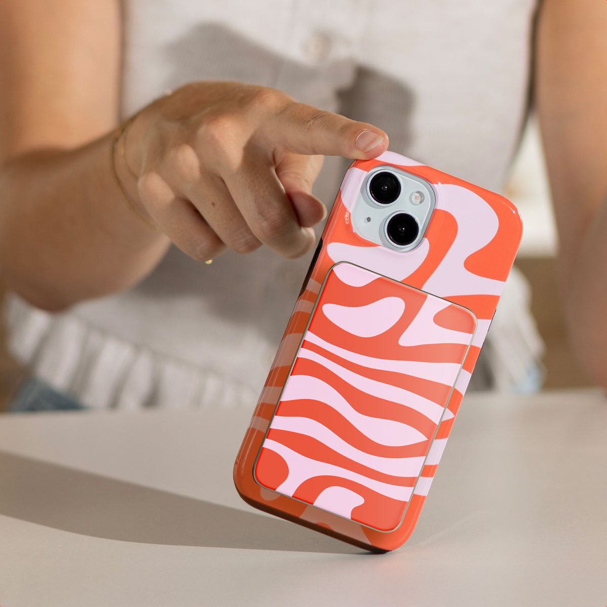 Een hand houdt een telefoon met een kleurrijke hoes vast. De hoes heeft een golvend oranje en roze patroon. De achtergrond is wazig.