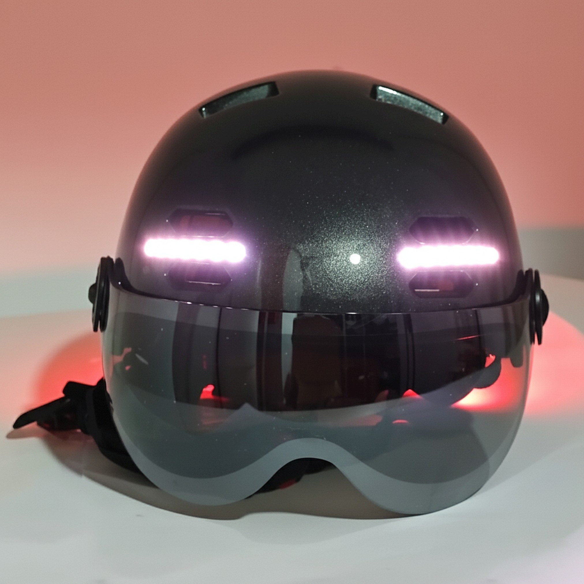 Ein dunkelgrauer Helm mit Visier und zwei leuchtenden Lichtern. Das Visier reflektiert das rote und weiße Umgebungslicht.